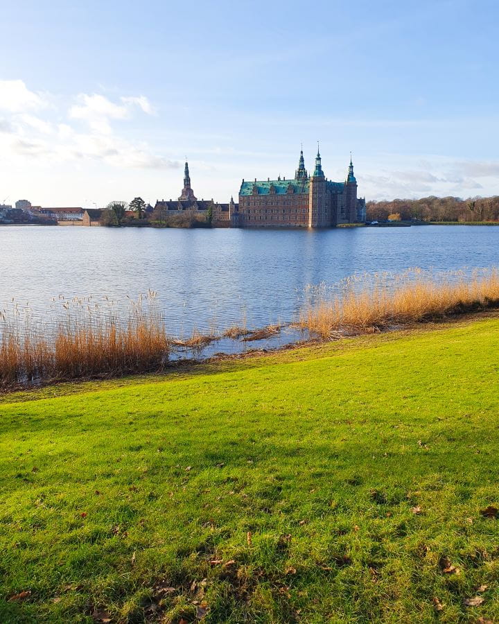 Denmark Explore Frederiksborg Castle In a Beautiful Day Trip from Copenhagen A stretch of green grass with taller brown grass on the edge of the water. There's a big blue pond behind it and on the other side is the big Frederiksborg castle in Denmark. It's brown with a green roof and there's four turrets coming off it. Explore Frederiksborg Castle In a Beautiful Day Trip from Copenhagen