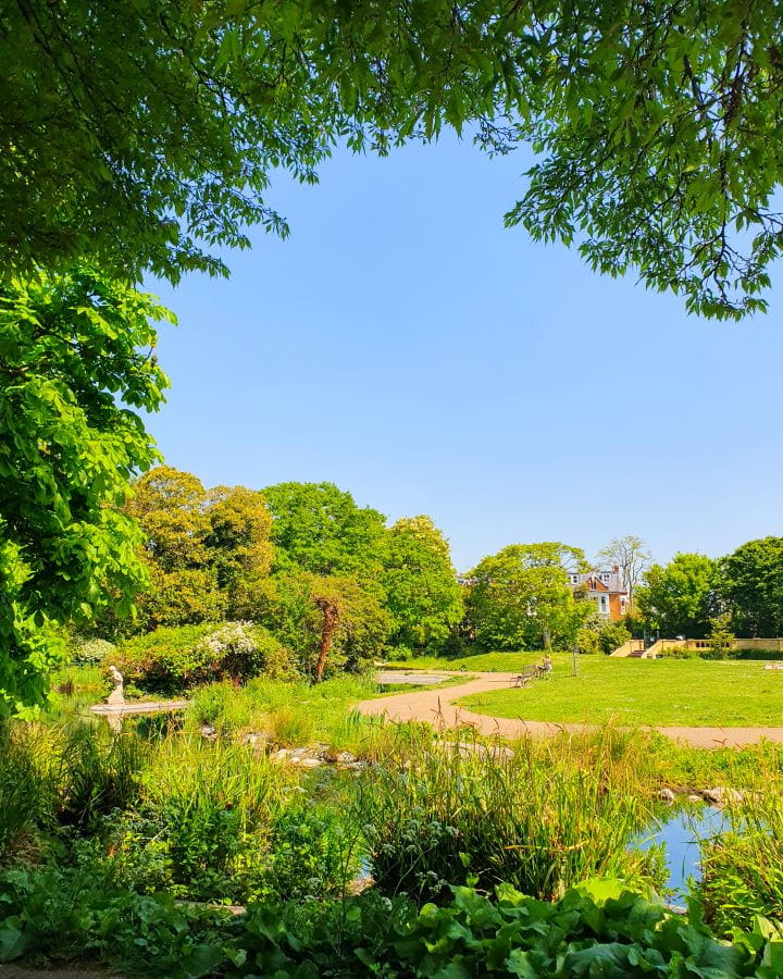 United Kingdom Bishop's Park Nine Less Touristy But Wonderful Parks to Visit in London There's green trees framing the photo, a pond is running through it with a path on the other side. There's green grass and trees behind the path in Bishop's Park, London. Nine Less Touristy But Wonderful Parks to Visit in London