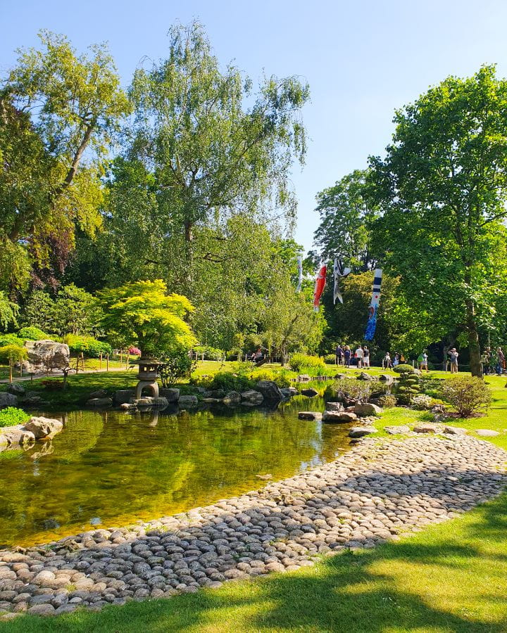 United Kingdom Kyoto Garden Holland Park Nine Less Touristy But Wonderful Parks to Visit in London Inside the Kyoto garden in Holland Park, London. There's small pebbles in the foreground with a pond behind it. There's a maple tree on the pond and a little statue, with more trees and flags behind it. There's also people walking around the pond. Nine Less Touristy But Wonderful Parks to Visit in London