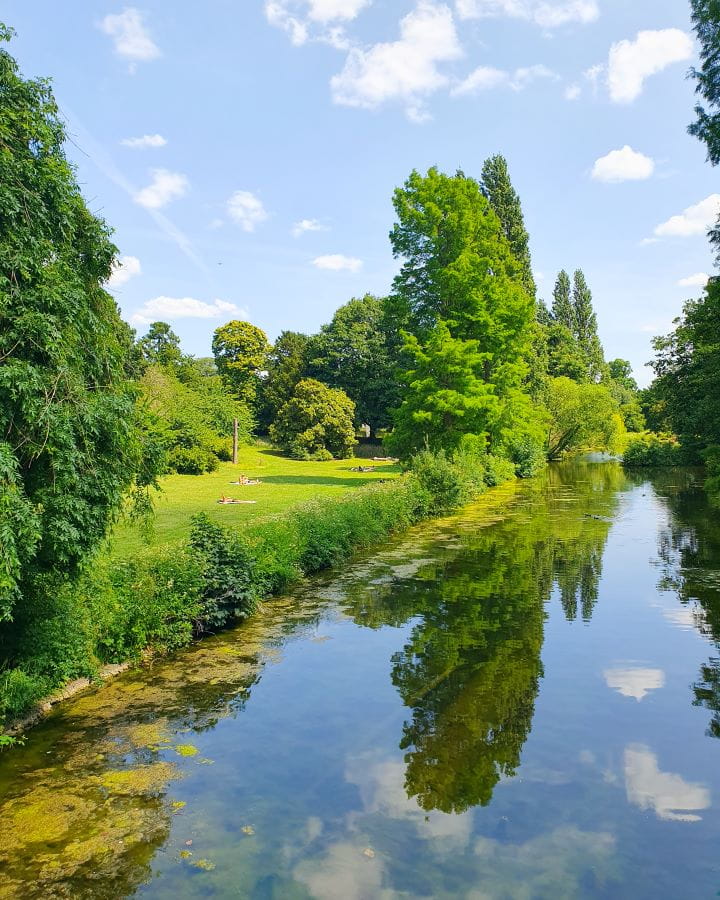 United Kingdom Chiswick house and gardens Nine Less Touristy But Wonderful Parks to Visit in London A pond in Chiswick house and garden in London. There's big trees on the other side of the pond and they're reflecting in the pond along with the blue sky above. There's more trees, green bushes and a big green open space with people picnicking on it. Nine Less Touristy But Wonderful Parks to Visit in London