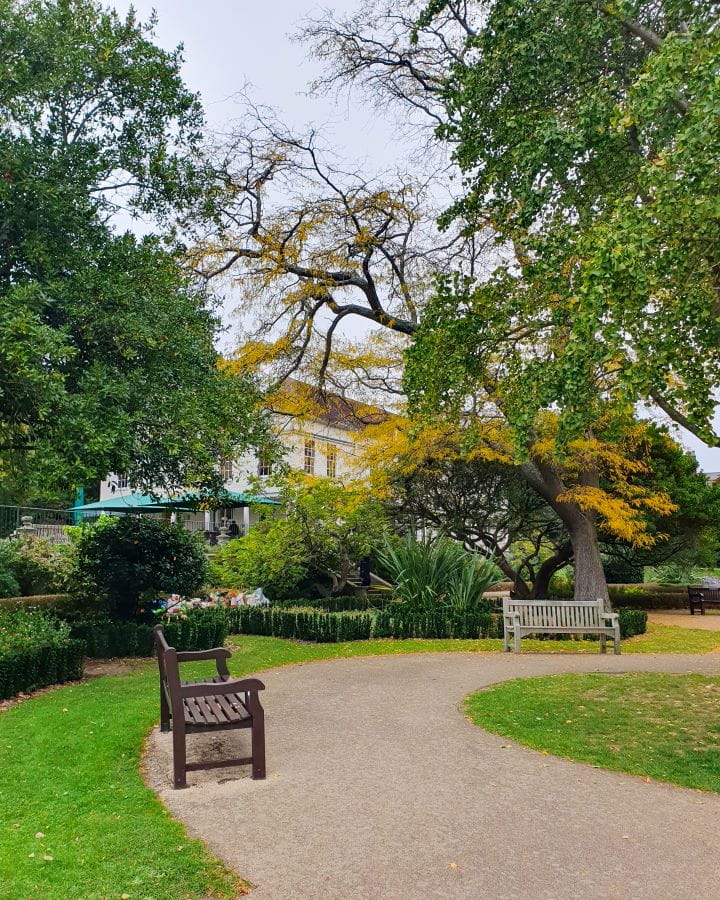 United Kingdom Waterlow Park Nine Less Touristy But Wonderful Parks to Visit in London A white path curving around with some wooden benches on it in Waterlow Park, London. There's green grass around the bath and lots of bushes and trees behind it. Peeking through those trees is Lauderdale house. Nine Less Touristy But Wonderful Parks to Visit in London