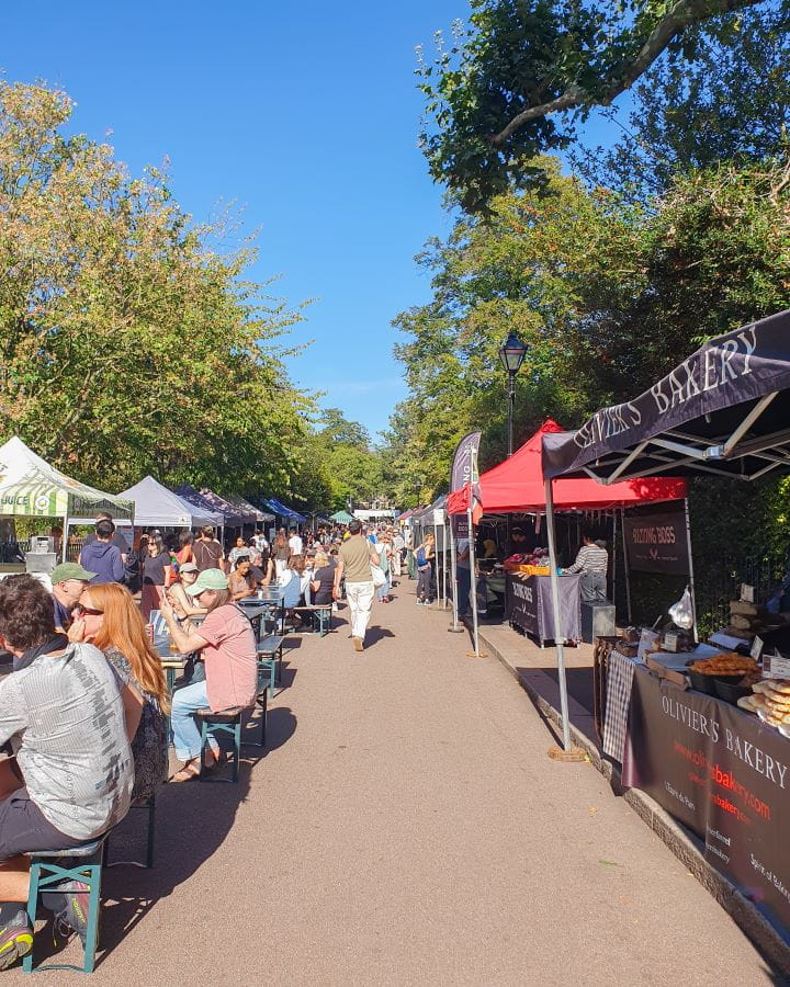 United Kingdom Victoria Park Nine Less Touristy But Wonderful Parks to Visit in London An outdoor market in Victoria Park, London. On the right of the path are market stalls in different colours selling bread and fruit. On the left are tables and chairs with people sitting at them and more market stalls behind them. There's also trees above the path and the sky is blue. Nine Less Touristy But Wonderful Parks to Visit in London